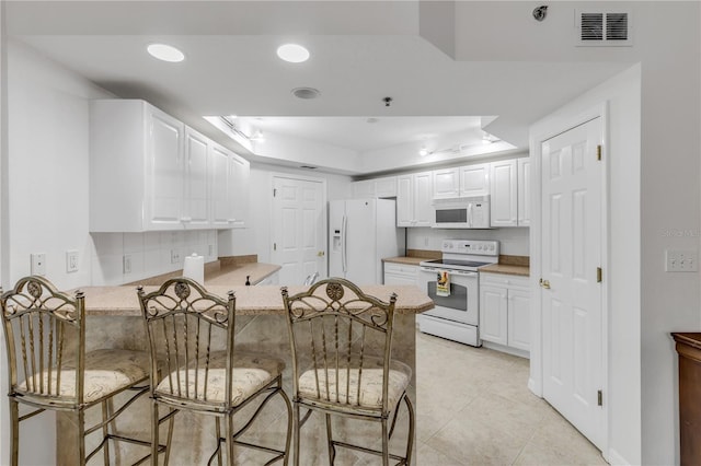 kitchen with white appliances, a raised ceiling, a peninsula, a breakfast bar, and white cabinets