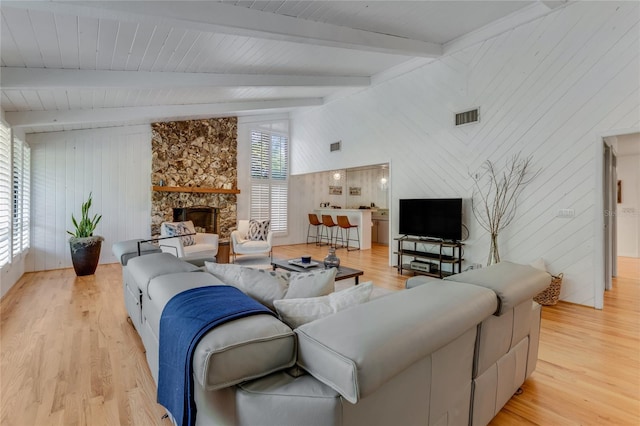 living room featuring lofted ceiling with beams, light wood-type flooring, visible vents, a stone fireplace, and wooden walls