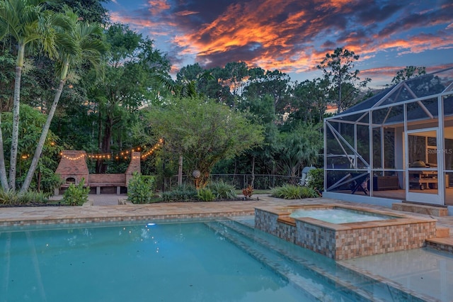 pool at dusk with an outdoor pool, an in-ground hot tub, glass enclosure, fence, and a patio area
