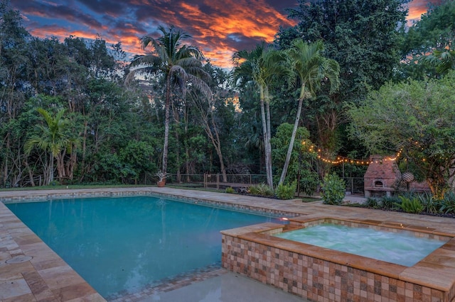 pool at dusk featuring exterior fireplace, an outdoor pool, fence, an in-ground hot tub, and a patio