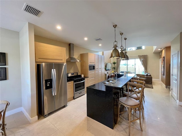 kitchen with an island with sink, light tile floors, stainless steel appliances, light brown cabinetry, and wall chimney exhaust hood