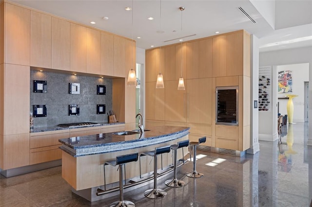 kitchen featuring light brown cabinetry, a kitchen island with sink, pendant lighting, dark stone countertops, and a breakfast bar