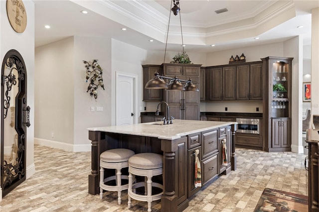 kitchen featuring light stone counters, a raised ceiling, dark brown cabinets, a breakfast bar, and a center island with sink