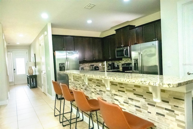 kitchen featuring light stone countertops, a kitchen breakfast bar, appliances with stainless steel finishes, dark brown cabinets, and tasteful backsplash