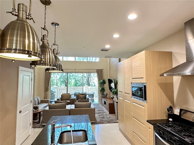 kitchen with stainless steel microwave, light tile floors, light brown cabinetry, stove, and wall chimney exhaust hood