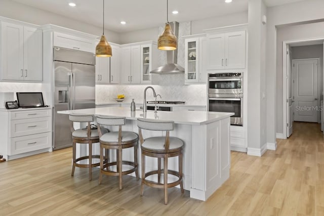 kitchen with pendant lighting, light hardwood / wood-style flooring, wall chimney exhaust hood, an island with sink, and appliances with stainless steel finishes