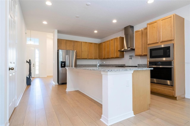 kitchen featuring appliances with stainless steel finishes, a center island with sink, light hardwood / wood-style floors, wall chimney range hood, and light stone counters