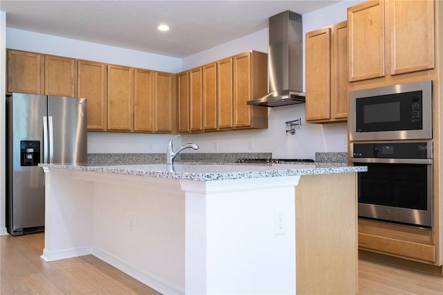 kitchen with light stone counters, appliances with stainless steel finishes, wall chimney range hood, and light wood-type flooring