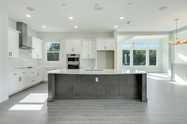 kitchen with wall chimney range hood, a wealth of natural light, an island with sink, and white cabinets