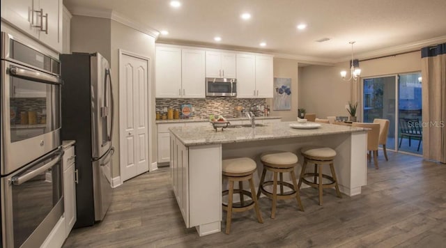 kitchen featuring appliances with stainless steel finishes, a kitchen island with sink, backsplash, white cabinetry, and decorative light fixtures