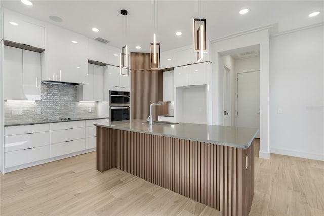 kitchen with stainless steel double oven, white cabinetry, sink, hanging light fixtures, and a center island with sink