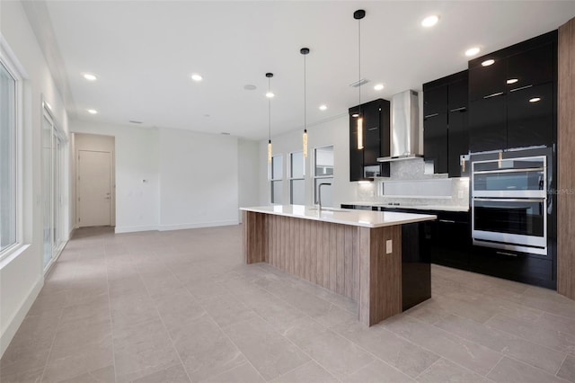 kitchen with decorative light fixtures, tasteful backsplash, an island with sink, sink, and wall chimney exhaust hood