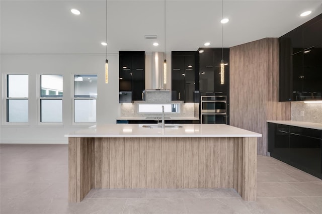 kitchen featuring sink, wall chimney range hood, a kitchen island with sink, tasteful backsplash, and stainless steel double oven