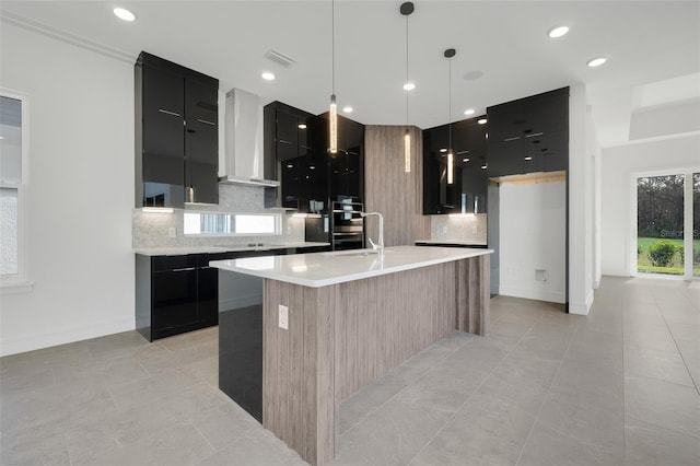 kitchen featuring a center island with sink, tasteful backsplash, wall chimney range hood, and decorative light fixtures