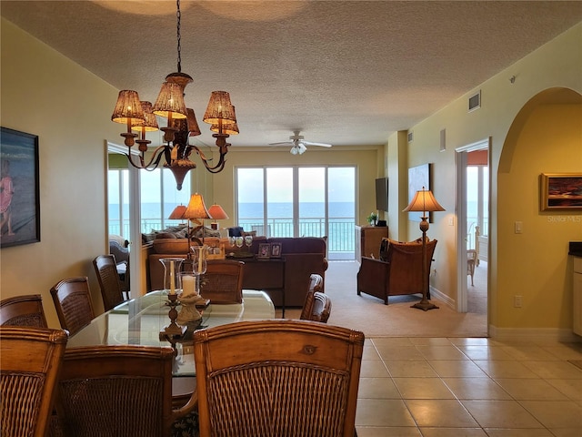 tiled dining space with a water view, a textured ceiling, and ceiling fan with notable chandelier