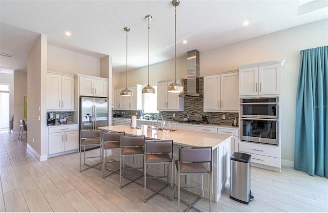 kitchen with a kitchen island with sink, backsplash, stainless steel appliances, wall chimney range hood, and decorative light fixtures