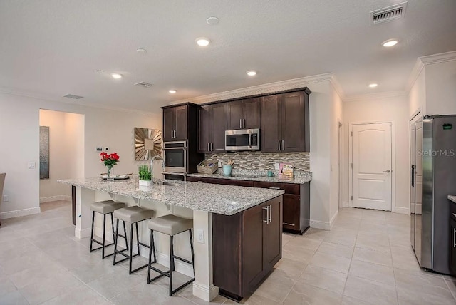 kitchen with dark brown cabinetry, a center island with sink, ornamental molding, stainless steel appliances, and decorative backsplash