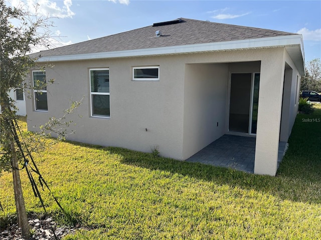 rear view of house featuring stucco siding, a lawn, roof with shingles, and a patio area