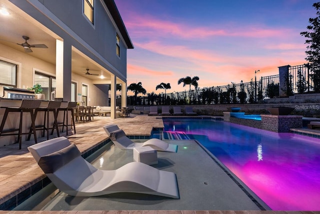 pool at dusk featuring a bar, ceiling fan, and a patio