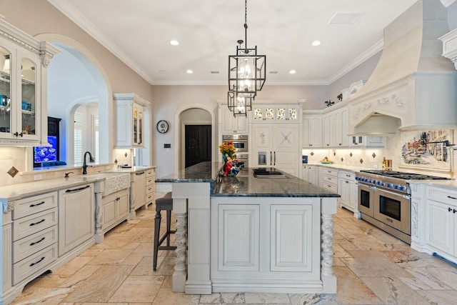 kitchen with a chandelier, custom range hood, a center island with sink, stainless steel appliances, and tasteful backsplash