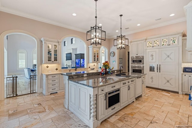 kitchen featuring light tile flooring, tasteful backsplash, stainless steel double oven, hanging light fixtures, and a kitchen island with sink