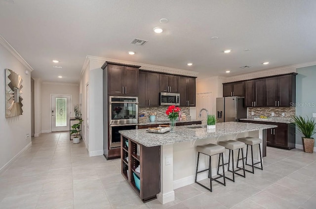 kitchen with light stone counters, dark brown cabinets, appliances with stainless steel finishes, and a center island with sink