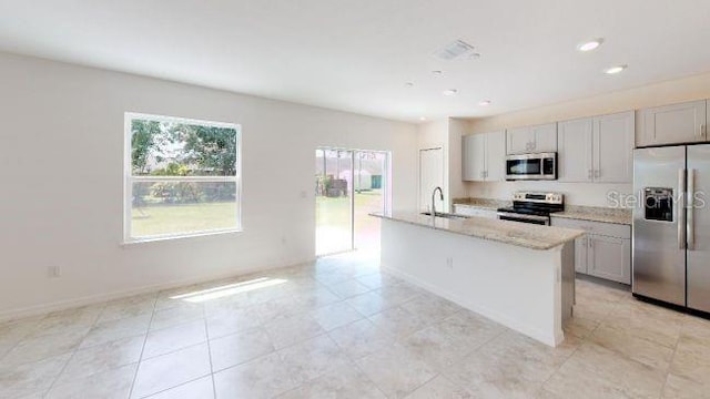 kitchen with light tile patterned flooring, appliances with stainless steel finishes, sink, light stone counters, and a center island with sink