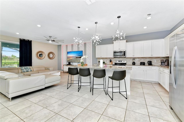 kitchen with a center island with sink, stainless steel appliances, light stone countertops, ceiling fan with notable chandelier, and white cabinetry