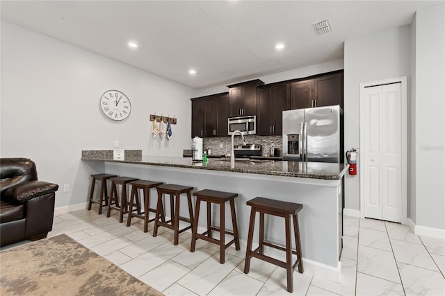 kitchen with kitchen peninsula, dark brown cabinets, backsplash, dark stone countertops, and appliances with stainless steel finishes