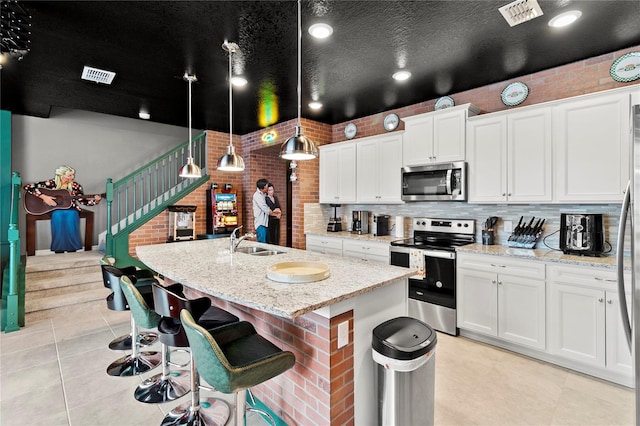 kitchen with pendant lighting, stainless steel appliances, brick wall, white cabinets, and a kitchen island with sink