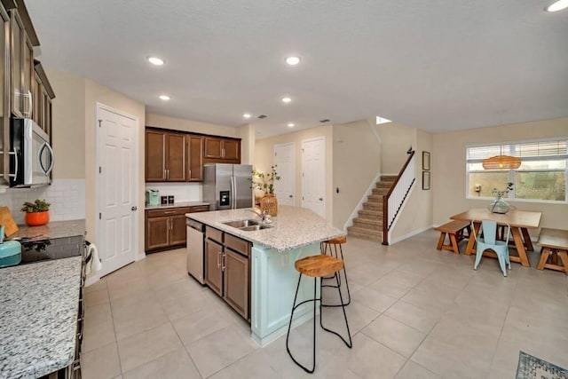 kitchen featuring an island with sink, sink, appliances with stainless steel finishes, light tile flooring, and light stone countertops