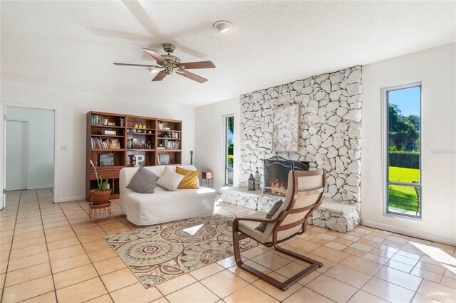 tiled living room featuring a textured ceiling, ceiling fan, a healthy amount of sunlight, and a stone fireplace