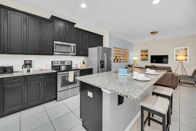 kitchen with light stone counters, light tile patterned floors, stainless steel appliances, a kitchen bar, and a center island with sink