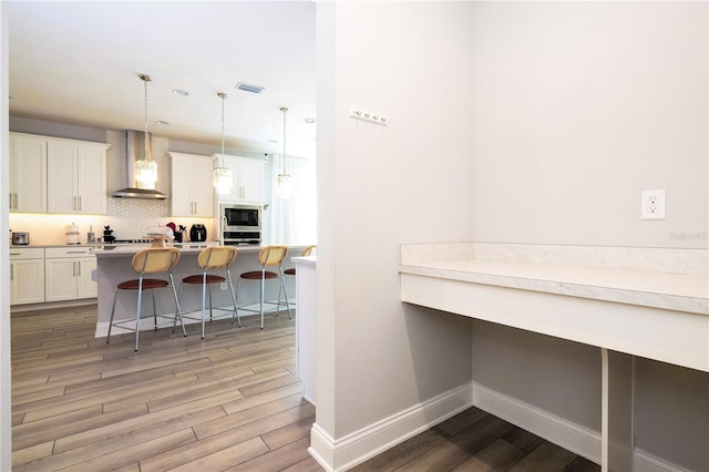 interior space with pendant lighting, a kitchen breakfast bar, stainless steel microwave, white cabinets, and wall chimney exhaust hood