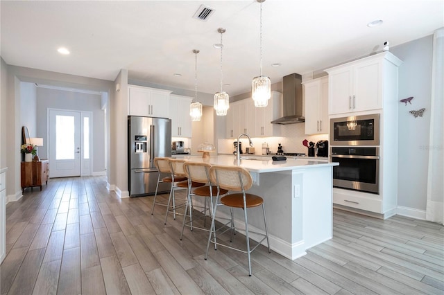 kitchen featuring wall chimney range hood, a center island with sink, white cabinets, light wood-type flooring, and stainless steel refrigerator with ice dispenser