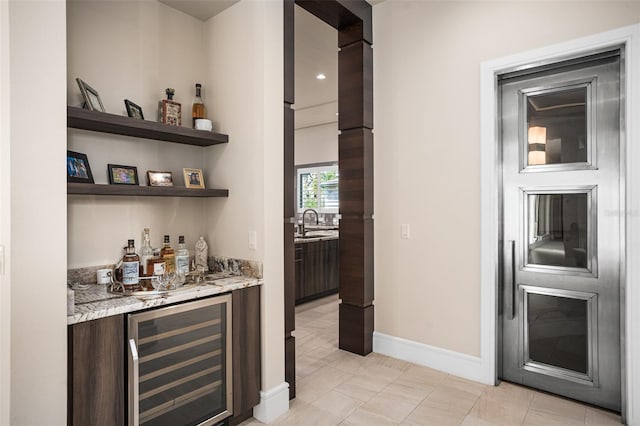 bar with wine cooler, dark brown cabinets, light stone counters, light tile patterned floors, and sink