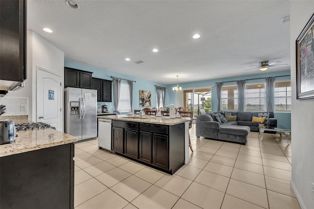 kitchen featuring decorative light fixtures, appliances with stainless steel finishes, an island with sink, light tile patterned floors, and light stone counters