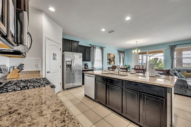 kitchen with light stone countertops, appliances with stainless steel finishes, decorative light fixtures, sink, and a kitchen island with sink