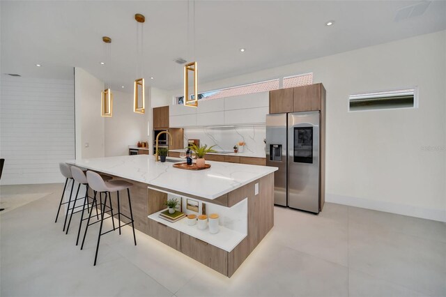 kitchen with an island with sink, stainless steel refrigerator with ice dispenser, hanging light fixtures, a kitchen breakfast bar, and backsplash