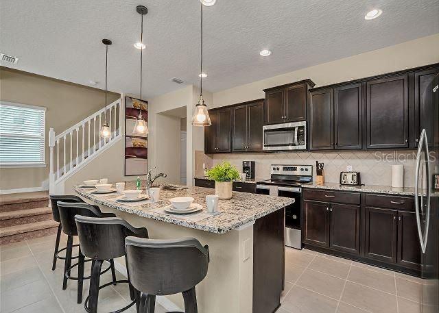 kitchen featuring hanging light fixtures, dark brown cabinets, stainless steel appliances, tasteful backsplash, and an island with sink