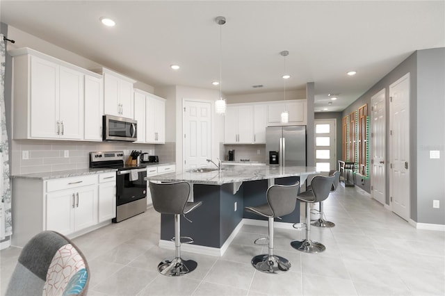 kitchen with an island with sink, pendant lighting, stainless steel appliances, light stone countertops, and white cabinets