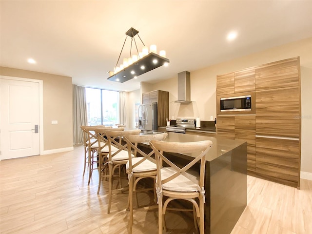 kitchen with stainless steel appliances, decorative light fixtures, wall chimney exhaust hood, light wood-type flooring, and sink