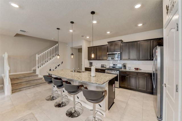 kitchen with hanging light fixtures, a kitchen island with sink, stainless steel appliances, light stone counters, and tasteful backsplash