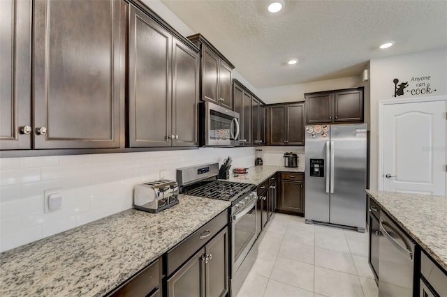 kitchen featuring dark brown cabinets, appliances with stainless steel finishes, light tile flooring, light stone countertops, and a textured ceiling