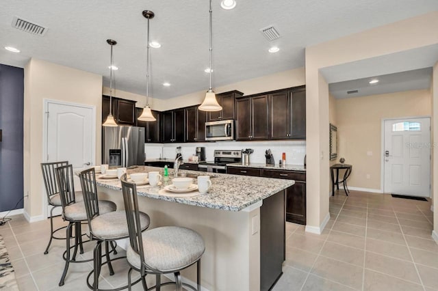 kitchen featuring a kitchen island with sink, pendant lighting, appliances with stainless steel finishes, light stone countertops, and a kitchen bar