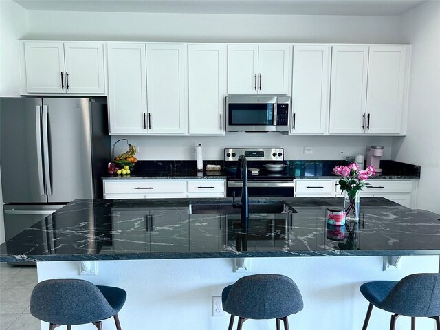 kitchen featuring white cabinetry, light tile flooring, stainless steel appliances, dark stone counters, and a breakfast bar area