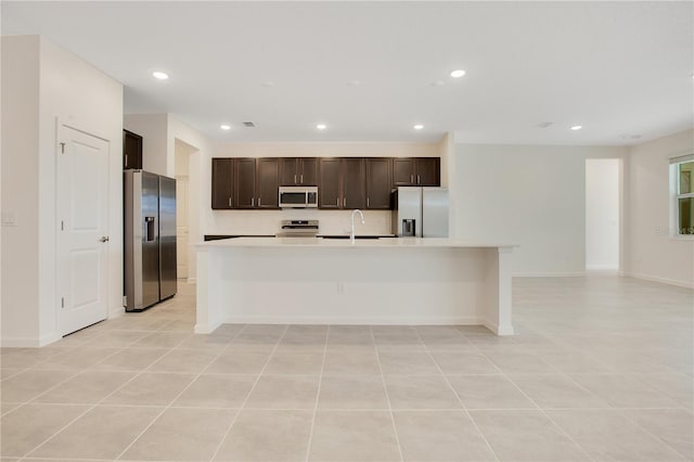 kitchen with stainless steel appliances, light tile flooring, dark brown cabinets, a kitchen island with sink, and sink