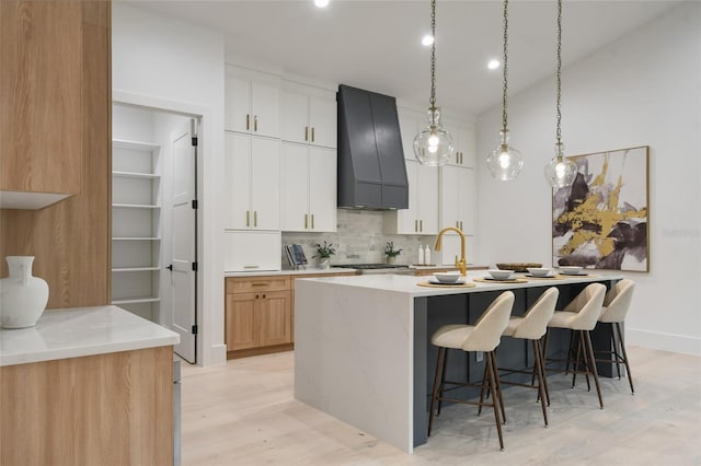 kitchen with white cabinets, custom range hood, light hardwood / wood-style flooring, and a kitchen island with sink