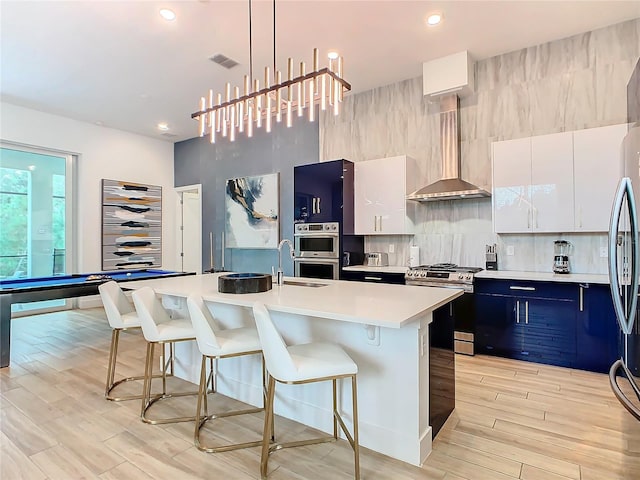 kitchen featuring wall chimney range hood, a kitchen bar, white cabinetry, appliances with stainless steel finishes, and a kitchen island with sink