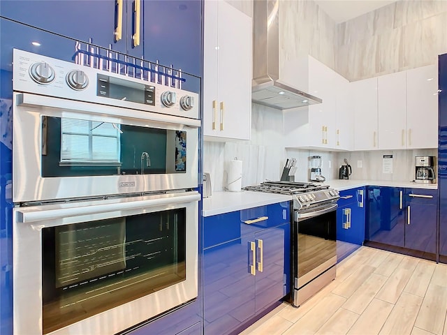 kitchen featuring white cabinetry, backsplash, blue cabinets, stainless steel appliances, and wall chimney range hood